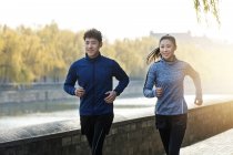 Smiling young asian man and woman jogging together near river — Stock Photo