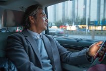 Maturo asiatico uomo d'affari utilizzando smartphone e guardando lontano in auto — Foto stock