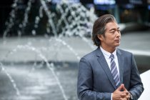 Serious mature asian businessman in formal wear standing near fountains — Stock Photo