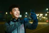 Улыбающийся азиат в наушниках и спортивной одежде делает селфи со смартфоном в ночном городе — стоковое фото