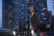 Mature asian man standing beside car and holding smartphone in night city — Stock Photo