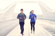 Вид счастливой молодой азиатской пары в спортивной одежде, бегущей по мосту и улыбающейся перед камерой — стоковое фото