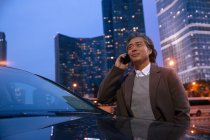 Улыбающийся взрослый азиат, стоящий рядом с машиной и разговаривающий по смартфону — стоковое фото