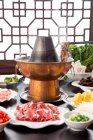 Pauzinhos com carne acima do cobre panela quente e pratos com vários ingredientes na mesa, conceito prato de atrito — Fotografia de Stock