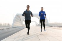 Спортивная молодая пара, улыбающаяся в камеру и бегающая вместе по мосту — стоковое фото