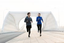 Full length view of happy sporty young asian couple smiling each other and running together on bridge — Stock Photo