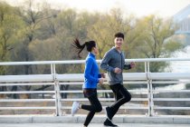 Lado vista de sporty joven asiático pareja sonriendo uno al otro y trotando juntos en puente - foto de stock