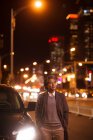 Maduro asiático hombre de pie al lado de coche y mirando lejos en noche ciudad - foto de stock