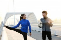 Счастливая молодая азиатская пара в спортивной одежде улыбаясь друг другу и растягиваясь вместе на мосту утром — стоковое фото