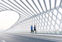 Ganzkörperansicht eines jungen Joggerpaares beim gemeinsamen Sportkleidungstraining auf einer modernen Brücke — Stockfoto
