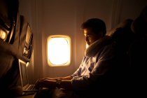Hombre joven usando el ordenador portátil mientras está sentado en el avión, vista lateral - foto de stock