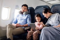 Família feliz com uma criança viajando de avião, pai dormindo e mãe com filha usando tablet digital — Fotografia de Stock