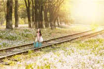Очаровательный азиатский ребенок в платье с сумкой на железной дороге возле цветочного поля — стоковое фото