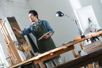 Фокусований чоловічий художник у фартусі, що тримає палітру та картини в студії, низький кут зору — стокове фото