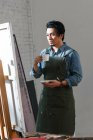Male asian painter drinking coffee and looking at easel with picture in studio — Stock Photo
