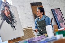 Concentrated male artist in apron looking at portrait on easel in studio — Stock Photo