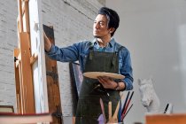 Focused male artist holding palette and painting picture in studio — Stock Photo