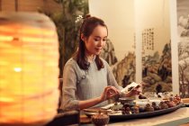 Красива усміхнена молода азіатка тримає сухе чайне листя над столом з посудом — стокове фото