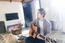 Guapo asiático hombre jugando acústica guitarra y mirando lejos en casa - foto de stock