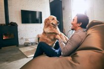 Vista lateral de feliz asiático homem sentado no feijão saco cadeira e brincando com o cão em casa — Fotografia de Stock