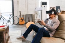 Emotionaler junger Mann in Virtual-Reality-Headset spielt zu Hause mit Joystick — Stockfoto