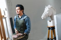 Ásia masculino artista no avental segurando paleta e pintura quadro no estúdio — Fotografia de Stock