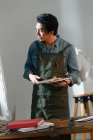Серйозний азіатський художник в фартусі тримає палітру і дивиться на картину в студії — стокове фото