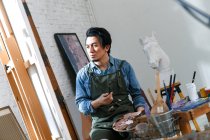 Serious asian male artist holding palette and looking at picture on easel in studio — Stock Photo