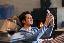 Smiling asian man sitting on bean bag chair and using smartphone at home — Stock Photo