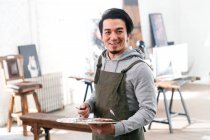 Handsome male painter holding palette and smiling at camera in studio — Stock Photo