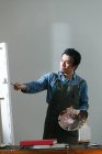 Красивий фокусований китайський художник в фартусі, що тримає палітру і картини в студії — стокове фото