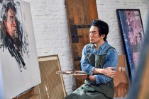 Серйозний чоловічий художник в фартусі тримає палітру і дивиться на портрет на мольберті в студії — стокове фото