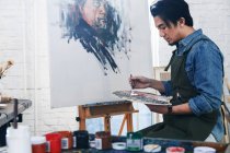 Handsome asian male artist in apron holding palette and painting portrait in studio — Stock Photo