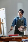 Bonito artista chinês no avental segurando paleta e pintura de imagem no estúdio — Fotografia de Stock