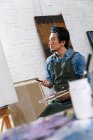 Вдумливий чоловічий художник в фартусі тримає палітру і дивиться на мольберт в студії — стокове фото