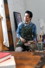 Серьезный азиатский художник в фартуке держа палитру и глядя на живопись в студии — стоковое фото