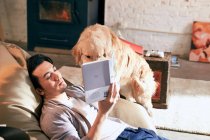 Aus der Vogelperspektive: junger asiatischer Mann liest Buch und spielt zu Hause mit Hund — Stockfoto
