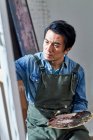 Concentrated male artist in apron holding palette and painting picture in studio — Stock Photo