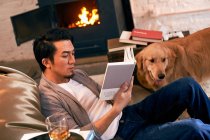 Vista lateral de centrado asiático hombre sentado en frijol bolsa silla y lectura libro, perro caminando al lado en casa - foto de stock
