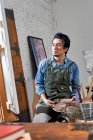 Серьезный азиатский художник в фартуке держа палитру и глядя на живопись в студии — стоковое фото