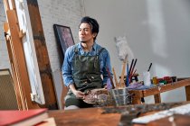 Сфокусированный художник в фартуке, держащий палитру и смотрящий на картину в студии — стоковое фото