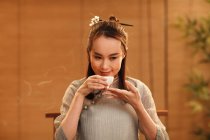 Beautiful smiling young chinese woman holding white cup and smelling aromatic tea — Stock Photo
