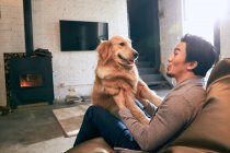 Lado vista de feliz asiático hombre sentado en frijol bolsa silla y jugando con perro en casa - foto de stock