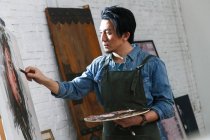Handsome asian man in apron holding palette and painting picture in art studio — Stock Photo