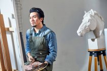 Pensive asian male artist holding palette and looking at picture on easel in studio — Stock Photo