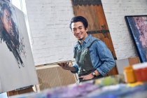Cheerful asian male artist holding palette and smiling at camera in studio — Stock Photo