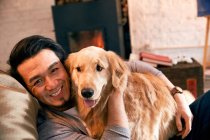Cheerful asian man resting with dog and smiling at camera at home — Stock Photo