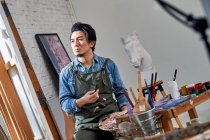 Low angle view of asian male artist in apron holding palette and painting picture in studio — Stock Photo