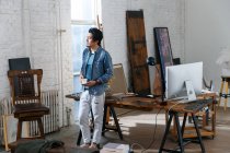 Pensativo asiático hombre celebración taza de café y mirando lejos en arte estudio - foto de stock