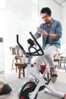 Smiling young asian man sitting on exercise bike and using smartphone at home — Stock Photo